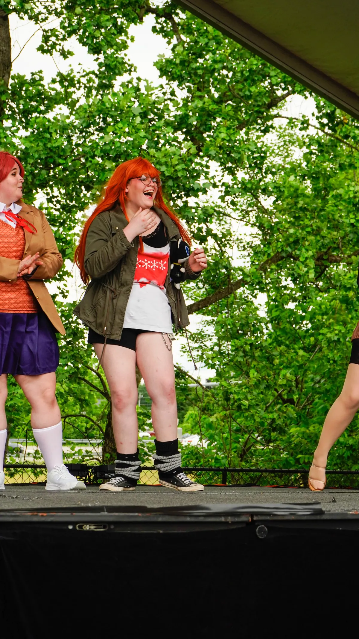 Two cosplayers stand on a tented outdoor stage, one in an orange wig laughing while another in a red wig and blazer claps beside her at Knox Pop Con.